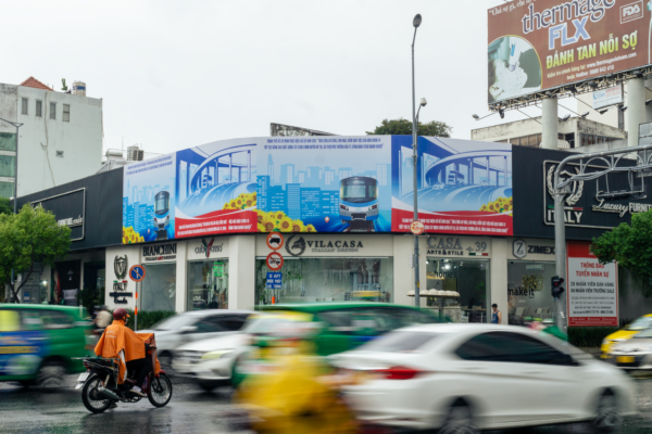 This landscape format photo shows a large, four-paneled propaganda sign overlooking a busy Ho Chi Minh City intersection through which taxis and scooters stream. The traffic appears blurred but the billboard and a lone scooter driver in an orange rain poncho appear sharp and crisp. The sky is cloudy. The billboard envelops a street corner building. The Vietnamese text urges safe adaptation to the COVID-19 pandemic and a continued push for economic growth. Two of the panels feature a digital image of the city's planned metro train in front of the city skyline. The other panels depict newly built highways.