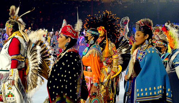A range of colorful indigenous regalia is worn by an intertribal group of men and women hopping rhythmically from right to left in profile. Colorful shawls with geometric embroidery and beaded headpieces, necklaces, and earrings give the women a dignified look. They seem serious and proud. The men are adorned with feathers in a range of styles and face painting, including a central figure with a bright yellow face topped with a red fringed and black feathered headdress. Some are wearing face masks that indicate caution at this early post-Covid event. The Eagle Staff that leads this procession can be seen in the distance.