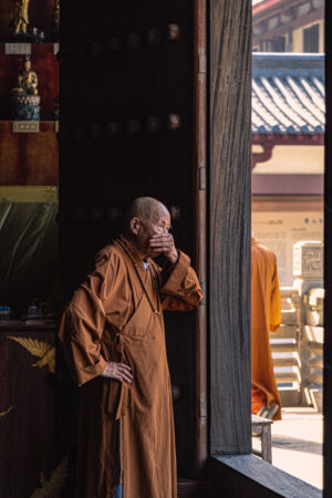A monk wearing an orange-brown cassock and Buddhist beads around his neck covers his mouth with his rough hands as he stands at the entrance of a doorway looking out. Faint door nails and glistening rows of enshrined Buddha statues can be seen in the background. On the right side of the picture, the back of a monk can be seen as he walks past the door.