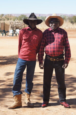 Two Australian Aboriginal men stand side by side wearing stockwork attire. The man to the left is wearing a black Akubra hat, red shirt, blue jeans and tan boots and is smiling to the camera with his hands behind his back. The man to the right is wearing a tan hat, sunglasses, a red checkered shirt, dark jeans and red boots and he is standing with his arms to his side. The background is of desert terrain with trees and mountains in the distance.