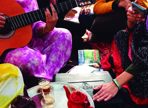 In the top left of the image a person wearing a pink lengthy fabric that wraps around the entire head and body strums a brown and black guitar. People sit next to them with various items in between them including a silver tray with glasses and tea pots on it, a black container, and a piece of green and blue glossy paper. To the right a girl photographs the gathering on her phone. 