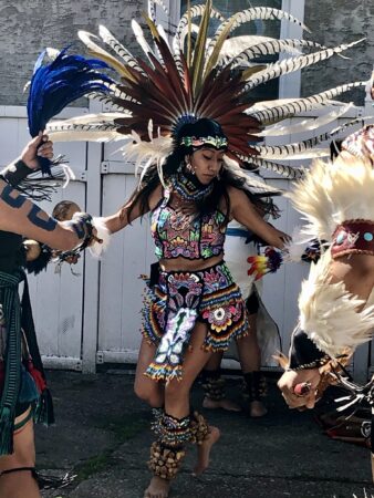 A woman in colorful, rainbow Aztec-inspired clothing and feather headdress dances in the center of the photo. The headdress is made of black-brown feathers at the center that become white with black stripes as it goes out. Two people frame her on either side of the image. To the left someone with tattooed arms holds a bunch of blue feathers. To the right is an arm covered in white feathers. The background features a white paneled door.