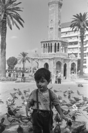 In this black and white photo, a small boy wearing a t-shirt and pants held up by overalls stands in front of several pigeons. In the background, a town square can be seen that features a clock tower framed by three palm trees. A building and several people can be seen in the distance.