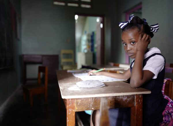 A young girl wearing a black and white school uniform sits in a dark classroom at school with only a small amount of light coming through the window. She is sitting propped up on one arm at a brown wooden desk and stares at the camera as she holds a pencil on the notebook with the other hand. The blackboard is in front of her desk. There are two big ribbons in her hair and her pink backpack hangs on the back of her chair. A few tables and chairs can be seen in the blurry background.