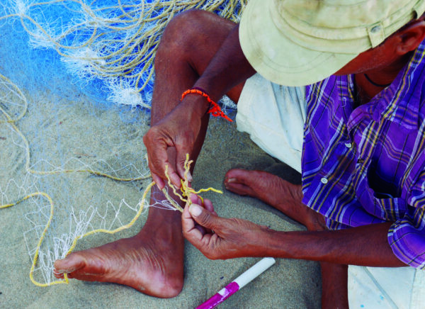 The close-up and top-down shot of the scene shows in detail the dexterity, skill and concentration of a Pagi fisherman mending a net while sitting on the golden sands of the Agonda beach in South Goa. He is wearing a stained tan hat, which covers his face, an orange bracelet, purple plaid shirt and khaki pants. His fingers deftly work through the white and tan threads, weaving and tying them together, while his bare foot holds one of the ropes that supports the web of threads. White, tan, and blue thread and netting lays next to him as he works.