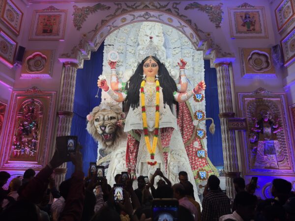 The center of a photo is dominated by a lit up Hindu shrine featuring a female Goddess statue with long black hair, four arms, and a white, red and gold dress. She is seated on a lion figure and holds various weapons, there is intricate white detailing in the back. Beside the shrine the walls feature detailed décor which is under darker lights with red and blue glowing lights highlighting it. A group of people can be seen below worshiping and taking photos of the shrine on their smartphones. The lighting highlights the shrine with the people being shown under a shadow.