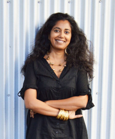 A picture of Mayanthi Fernando standing outside smiling against a white wall