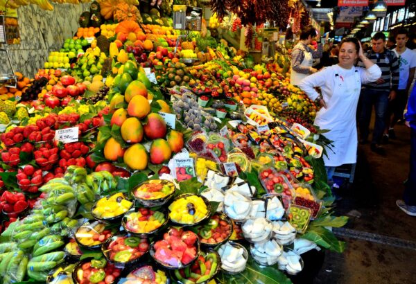 A woman wearing a white apron stands smiling, next to a market display of different fruits and vegetables.